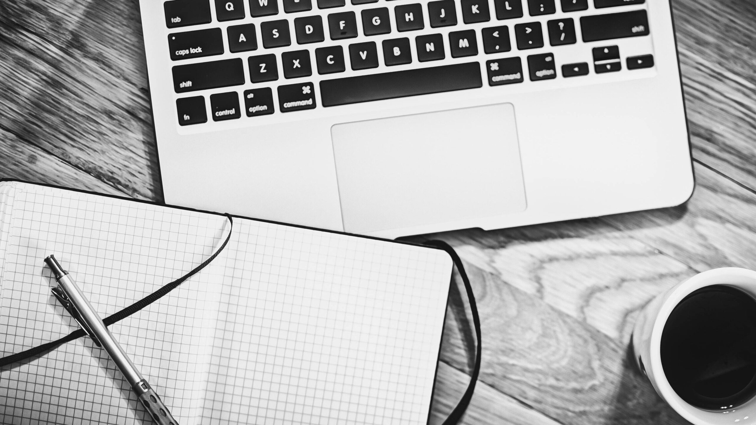Black and white image of a workspace featuring a laptop, notebook, pen, and coffee cup.